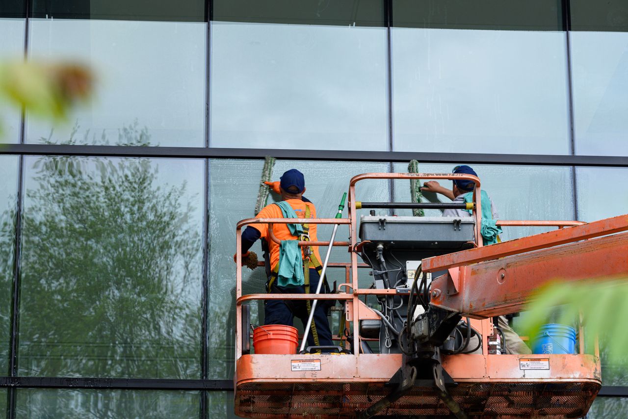 about-us Two workers cleaning windows on a glass building facade using a lift platform, enhancing building aesthetics.